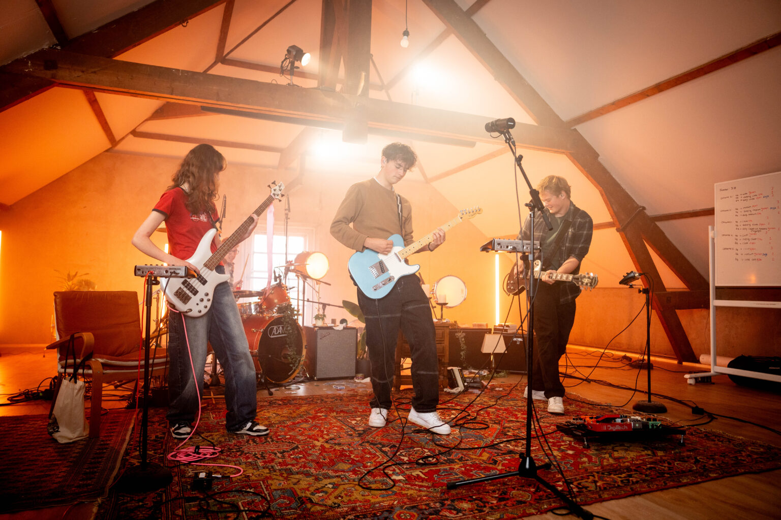 Drie jonge muzikanten spelen bas en gitaar op een zolder met houten balken, warm licht en een rood tapijt